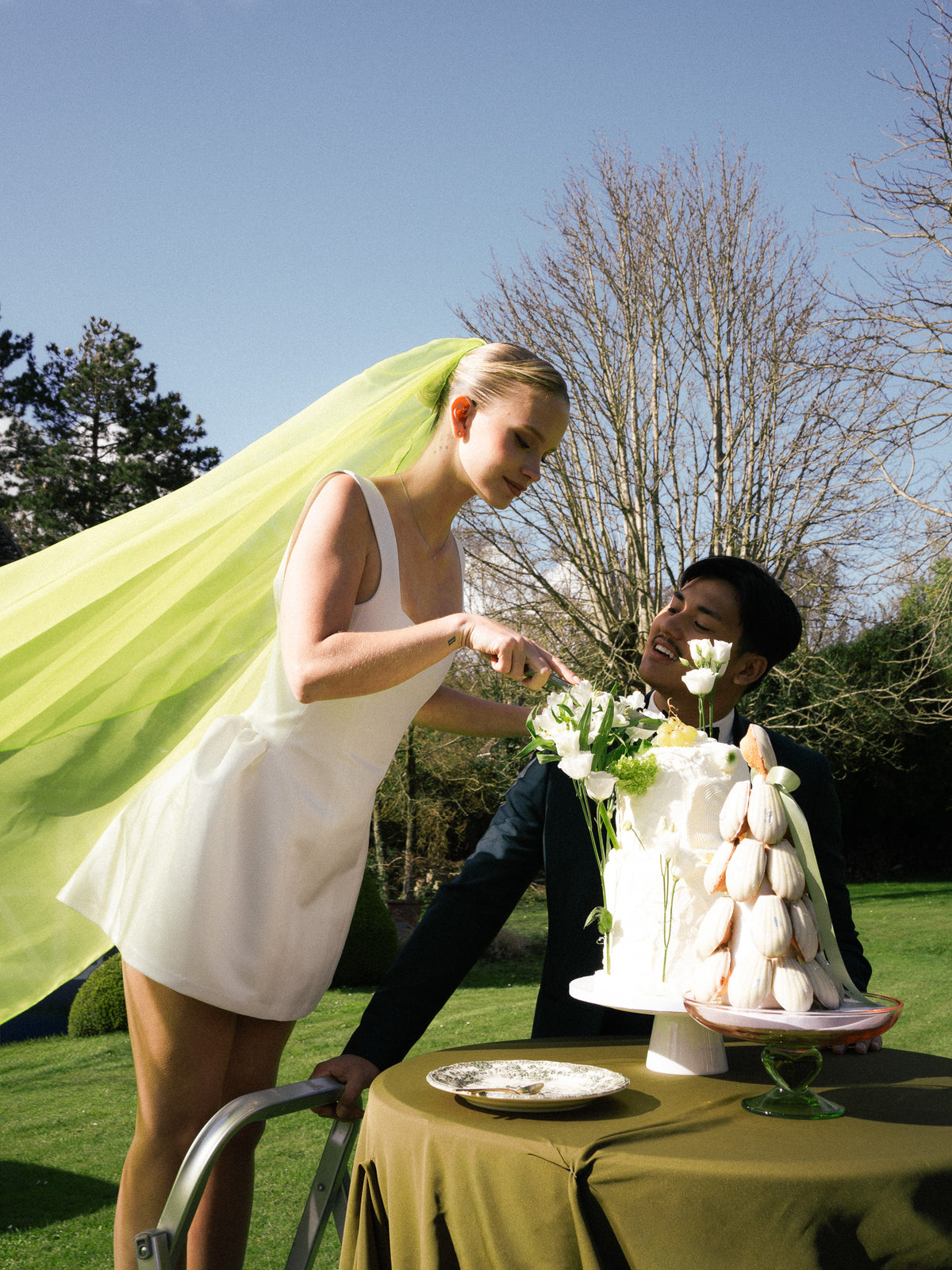 Mariée en robe courte blanche et voile vert chartreuse CANEZAN, coupant le gâteau de mariage avec le marié dans un jardin